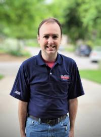 A man stands outside on a sidewalk, wearing a navy blue polo shirt with greenery and blurred cars in the background.