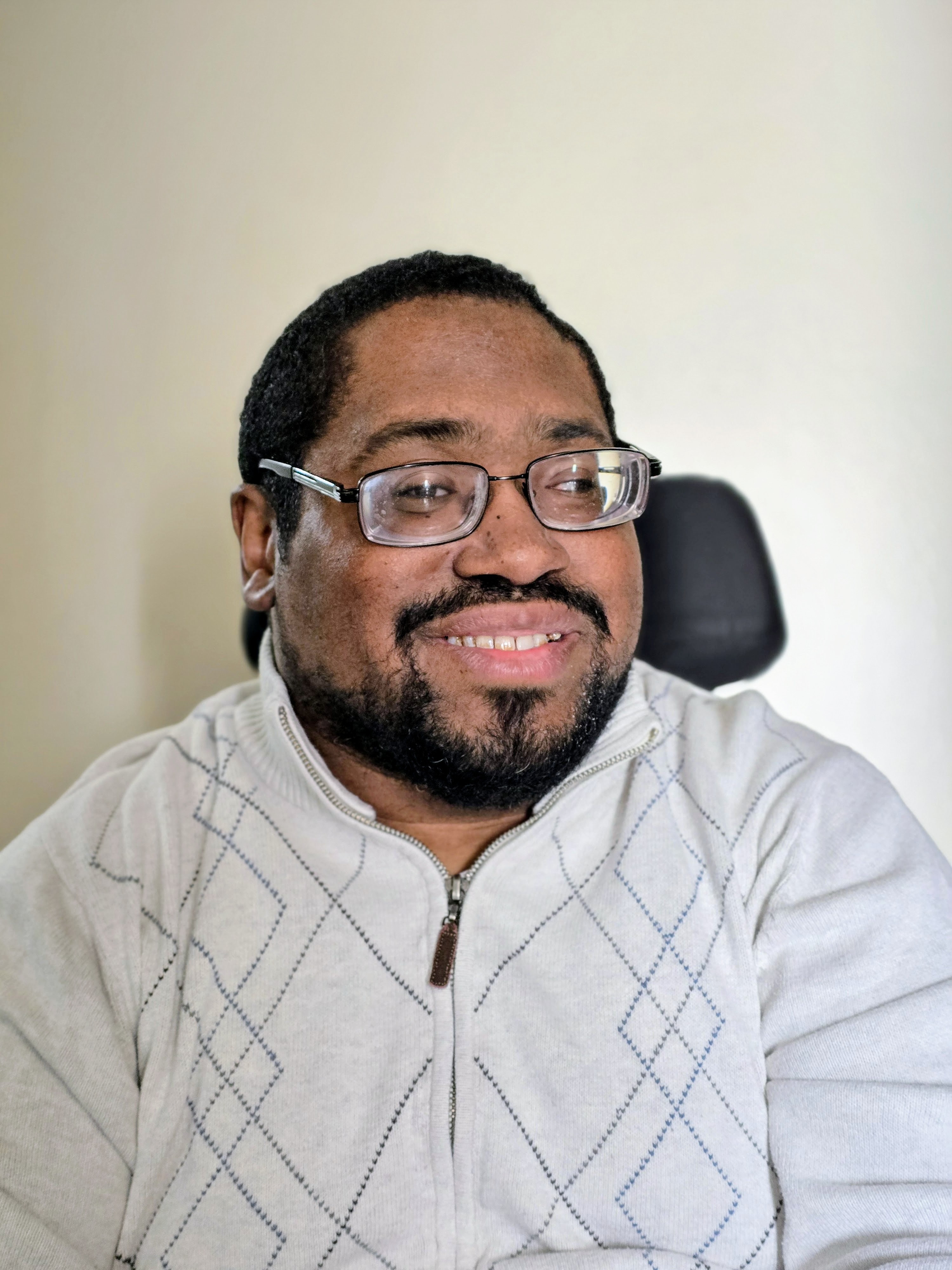 a Black man with a beard wearing glasses sitting. he is wearing a light-colored zip-up sweater